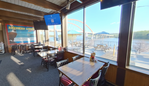 Interior of a restaurant with tables, large windows showing a river and bridge, and outdoor seating under umbrellas.
