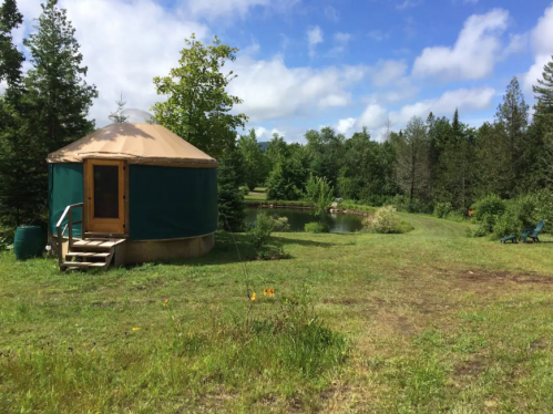 A green yurt sits in a grassy area near a pond, surrounded by trees and blue skies with scattered clouds.