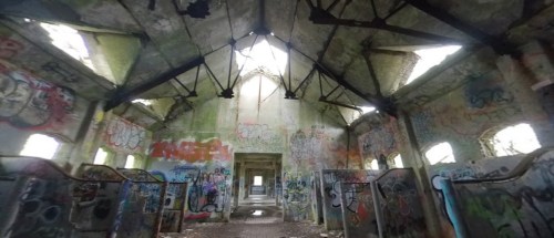 Interior of an abandoned building, featuring graffiti-covered walls and a dilapidated roof with light streaming through.