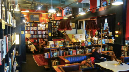 Cozy bookstore interior with bookshelves, seating areas, and warm lighting; people reading and a dog resting.