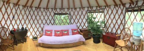 Cozy yurt interior with wooden furniture, a sofa with decorative pillows, plants, and a water cooler. Natural light from windows.