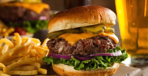 A juicy cheeseburger with lettuce, pickles, and onions, served with fries and a glass of beer in the background.