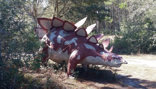 A life-sized dinosaur sculpture resembling a stegosaurus, surrounded by trees and shrubs in a natural setting.