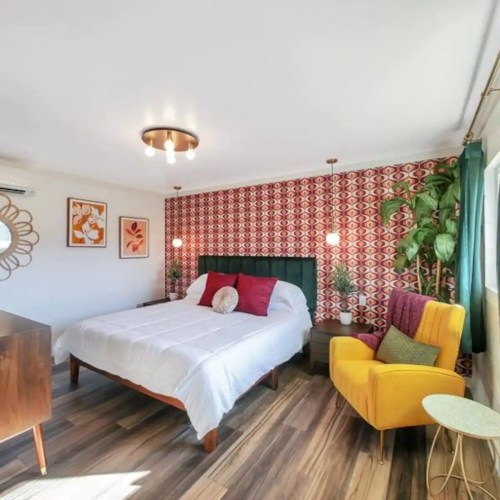 A stylish bedroom featuring a bed with colorful pillows, patterned wallpaper, a mirror, and a cozy seating area.