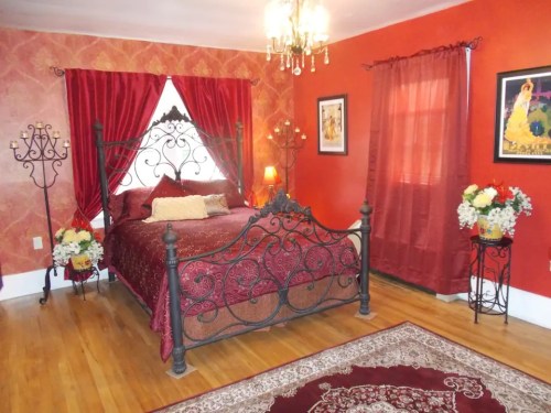 Cozy bedroom with a wrought-iron bed, red walls, elegant curtains, and decorative flowers. Warm lighting adds charm.