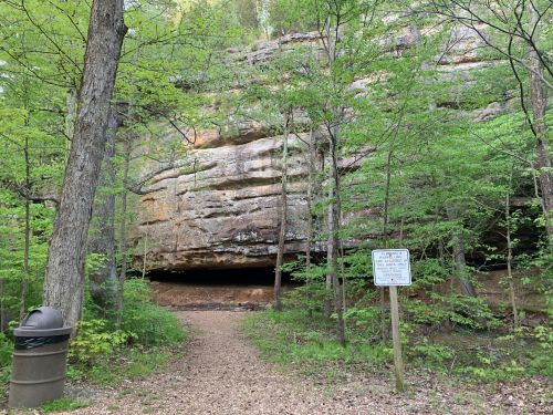 A forested area with a large rock overhang and a sign near a gravel path, surrounded by green trees.