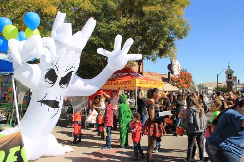 A large inflatable ghost towers over a busy street festival with families and vendors celebrating Halloween.