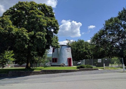 A quirky coffee pot-shaped building surrounded by trees and blue skies, with a gravel path in front.