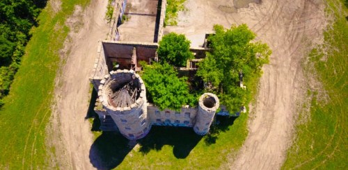 Aerial view of a partially ruined castle surrounded by greenery and dirt paths.