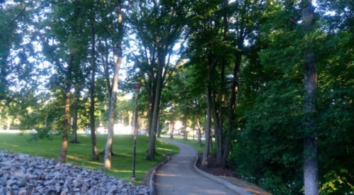 A winding path through a lush green park, lined with trees and a stone wall on one side.