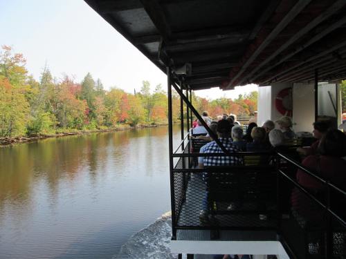 A boat filled with passengers glides along a calm river, surrounded by trees displaying autumn colors.