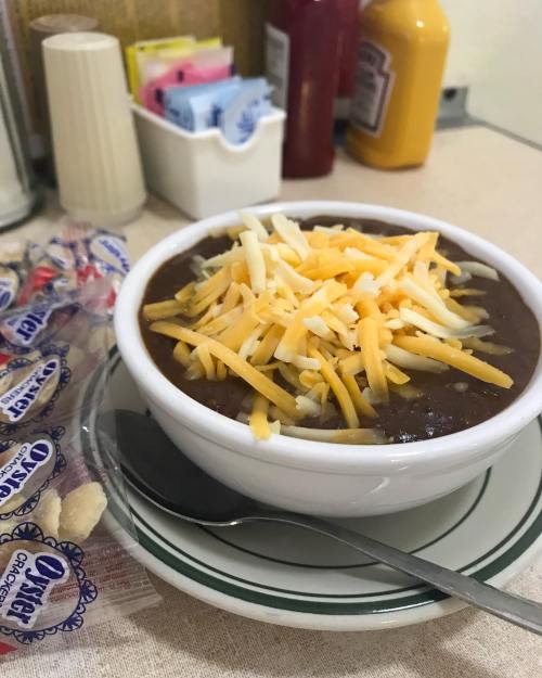 A bowl of chili topped with shredded cheese, next to a packet of oyster crackers on a table.