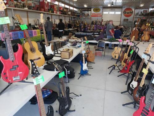 A music store interior filled with various guitars on display, with shoppers browsing and tables set up.