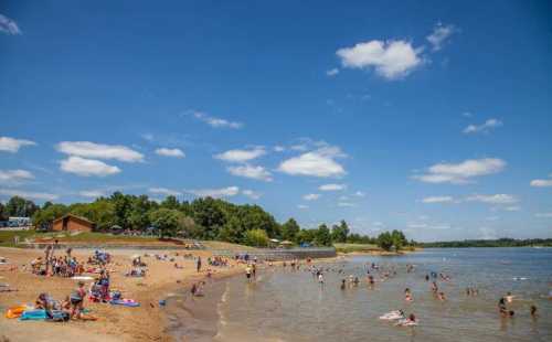 A sunny beach scene with people swimming and relaxing by the water under a blue sky with fluffy clouds.