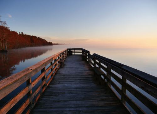 A wooden pier extends into a calm lake at sunrise, surrounded by trees with autumn foliage.