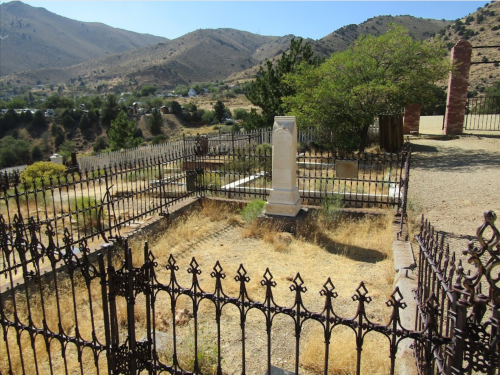 A weathered graveyard with a white tombstone, surrounded by a wrought iron fence and dry grass, set against a mountainous backdrop.
