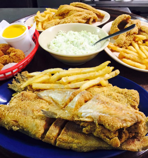 A platter of fried fish with fries, coleslaw, hushpuppies, and a bowl of dipping sauce on a wooden table.