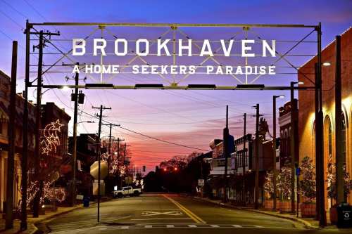 Sign reading "Brookhaven: A Home Seekers Paradise" at sunset, with decorated buildings and streetlights in view.
