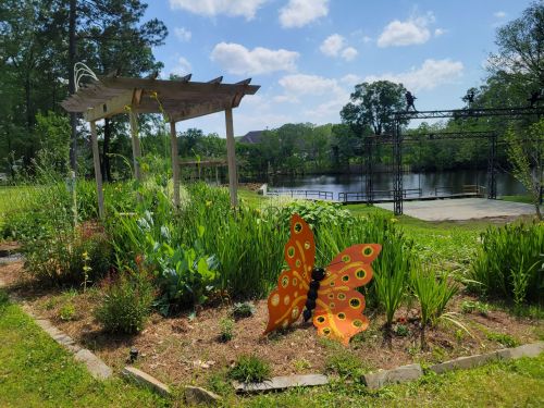 A vibrant garden with a large orange butterfly sculpture, green plants, and a serene lake in the background under a blue sky.