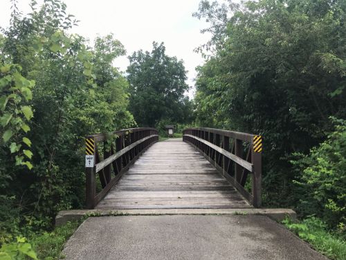 A wooden bridge spans a path surrounded by lush greenery and trees, leading into a natural area.