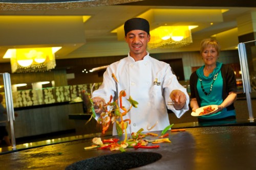 A chef in a white coat tosses colorful vegetables in a cooking station, while a woman watches nearby.