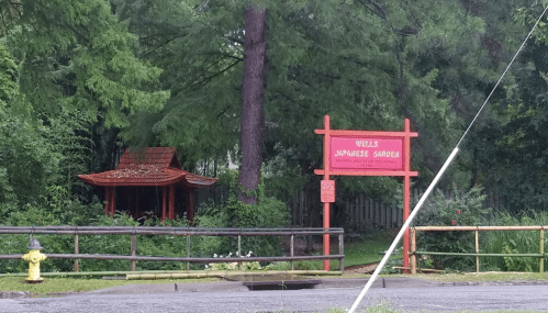 A red sign reading "Wells Japanese Garden" near a traditional gazebo surrounded by lush greenery.