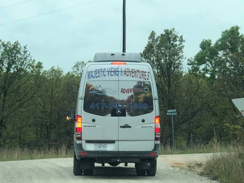 A white van with "Majestic Views & Adventures" written on the back, driving on a gravel road surrounded by trees.