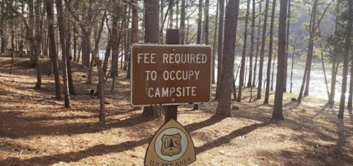 A sign in a wooded area indicating a fee is required to occupy the campsite, with a river visible in the background.