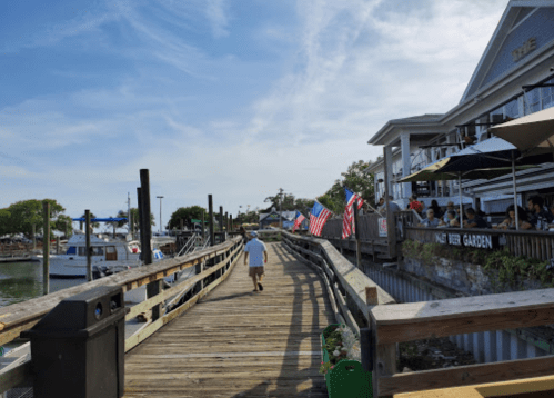 A child walks along a wooden pier lined with flags, near a waterfront restaurant and boats in the background.