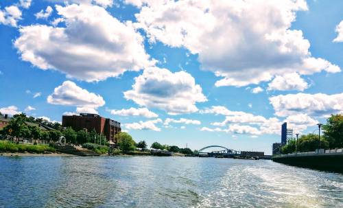 A scenic view of a river with fluffy clouds, greenery, and buildings along the shore under a bright blue sky.