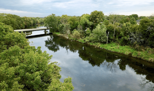 A serene river scene with lush greenery and a bridge crossing over calm waters under a cloudy sky.