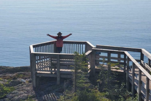 A person stands with arms outstretched on a wooden lookout platform by the calm blue sea.