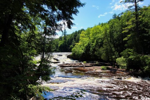 A serene river flows through a lush green forest under a bright blue sky, with two people standing on the rocks.