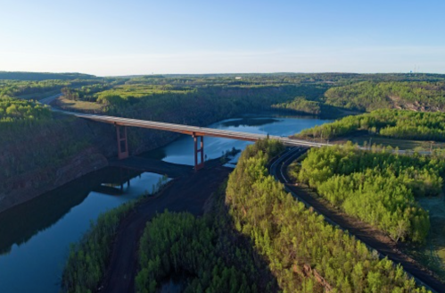 Aerial view of a bridge spanning a river, surrounded by lush green forests and hills under a clear blue sky.