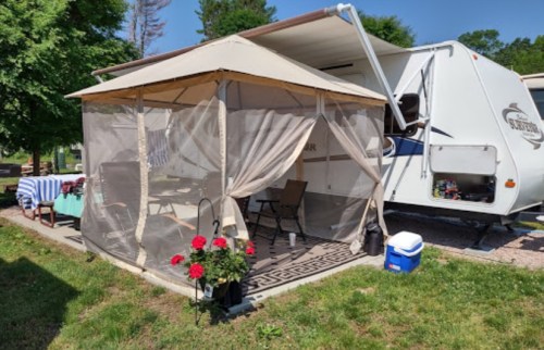 A screened tent with chairs and flowers beside a parked RV in a sunny outdoor setting.