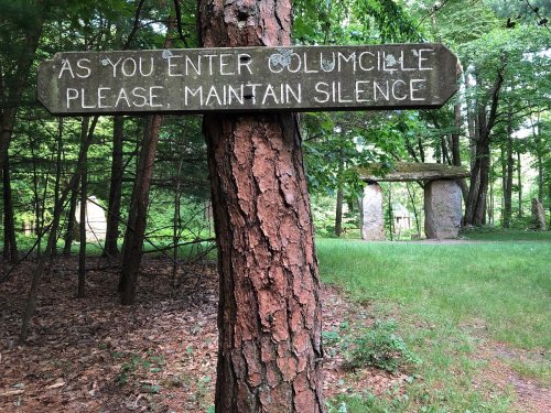 A wooden sign on a tree reads, "As you enter Columcille, please maintain silence," near stone structures in a forest.