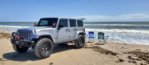 A silver Jeep Rubicon parked on a sandy beach near the ocean, with two folding chairs set up nearby.