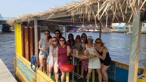 A group of ten people on a boat with a thatched roof, enjoying drinks by the water on a sunny day.