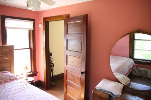 Cozy bedroom with a pink wall, wooden door, mirror, and a bed with patterned bedding. Natural light from the window.