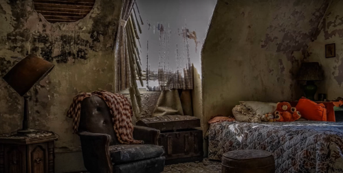 A dimly lit, worn bedroom with peeling walls, a cozy bed, an armchair, and a window with tattered curtains.