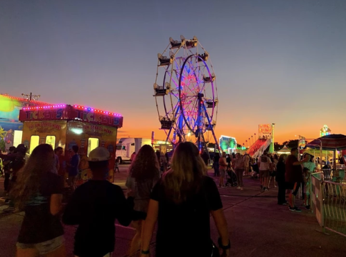 A vibrant carnival scene at sunset, featuring a Ferris wheel and colorful lights, with people walking through the fair.