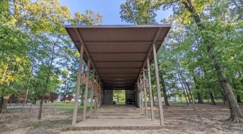 A spacious, open pavilion surrounded by trees, featuring a metal roof and concrete floor.