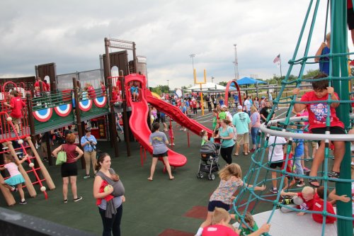 A busy playground with children playing on slides and climbing structures, surrounded by families and festive decorations.