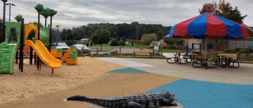 A colorful playground with slides, a picnic area, and a crocodile sculpture on a rubber surface. Trees in the background.