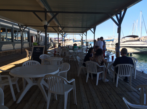 Outdoor dining area by the water, featuring white tables and chairs, with boats docked in the background.