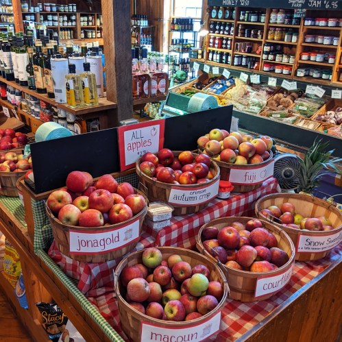 A rustic market display featuring various apple varieties in baskets, surrounded by shelves of sauces and preserves.