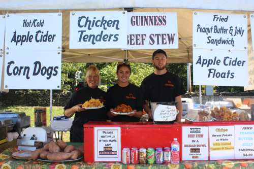 A food stall with three staff members holding plates of food, featuring signs for various menu items like chicken tenders and apple cider.