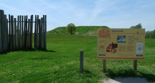 A grassy landscape featuring a wooden stockade and an informational sign about a historical site.
