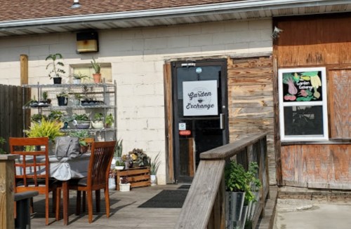 A cozy garden shop entrance with outdoor seating and plants displayed on shelves.
