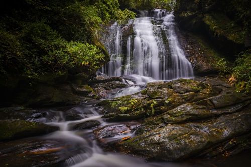 A serene waterfall cascades over rocky terrain, surrounded by lush greenery and soft flowing water.